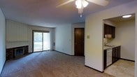 This single-floor residence features an open-concept living area finished with wall-to-wall carpeting and neutral-toned walls that lend a clean, understated aesthetic throughout. A ceiling fan with integrated light fixture serves as the central overhead lighting source in the main living space, promoting air circulation and comfort. A large sliding glass door dominates one wall of the living area, inviting natural light deep into the interior while providing direct access to an outdoor patio area, where views of surrounding greenery are visible. A floor-mounted wall vent indicates a built-in climate control system. A solid wood interior door provides access to an additional room, adding functional separation within the layout. Transitioning from the living area, a partial wall division leads into a connected kitchen space, where dark cabinetry and a sink are visible, suggesting a practical galley-style configuration. The overall layout is straightforward and functional, with an efficient use of space that connects the main living and kitchen areas seamlessly while maintaining distinct zones within the single-floor footprint.