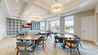 Located at 1231 Arya Way in Ocoee, Florida, this single-floor community space is thoughtfully designed to serve as a welcoming and functional gathering hub. The interior showcases a contemporary aesthetic defined by coffered ceilings, large pendant light fixtures, and light wood-style flooring that flows seamlessly throughout.

The space is divided into several distinct functional zones. A generously sized dining area features multiple square tables paired with upholstered armchairs in soft blue and gray tones, creating a comfortable setting for casual meals or social gatherings. Nearby, a dedicated lounge area offers plush sofas and accent chairs arranged around a glass-top coffee table and a mounted flat-screen television, all set against a dramatic wall of built-in open shelving. A linear fireplace adds warmth and visual interest to the space.

A business center alcove provides workstations equipped with computers and additional shelving, accommodating residents or visitors who need a productive workspace. Large windows throughout the space invite an abundance of natural light and offer views of the surrounding outdoor landscape, including palm trees and exterior signage. The overall ambiance is bright, modern, and inviting, balancing professional functionality with comfortable, resort-style design.