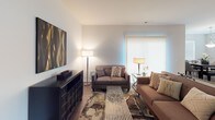 This single-floor residence offers a thoughtfully designed layout with modern finishes and a warm, neutral palette throughout. The kitchen is a standout feature, showcasing dark espresso cabinetry, granite countertops, stainless steel appliances including a gas range, microwave, and refrigerator, and elegant pendant lighting over a peninsula with a built-in sink. Light hardwood-style flooring ties the kitchen seamlessly into the adjacent spaces. The dining area benefits from an abundance of natural light streaming through multiple large windows dressed with horizontal blinds, and is anchored by a sophisticated chandelier above a dark wood dining table. The living room offers a comfortable, open atmosphere with hardwood-style flooring, vertical blinds filtering soft light, and ample space for seating. The primary bedroom is generously sized with plush carpeting, large windows framing outdoor greenery, and a serene, restful ambiance. A second bedroom comfortably accommodates two beds, also with soft carpet and good natural light. The bathrooms are cleanly finished with wood-look flooring, chrome fixtures, and glass-door shower enclosures. Throughout the space, a consistent modern aesthetic is maintained with crisp white walls, clean architectural lines, and well-considered details.