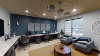 Located in Spring, Texas, this single-floor commercial office space combines functional design with refined aesthetics. The interior is anchored by a bold navy accent wall that serves as a striking backdrop for a long, multi-workstation desk equipped with multiple computer monitors and a multifunction printer — ideal for staff or resident use. A set of four decorative mosaic-style wall panels adds an artistic focal point to the workspace.

The lounge area offers a welcoming atmosphere with two upholstered blue accent chairs, coordinating throw pillows, and a round wood-and-metal coffee table set atop a woven jute area rug. Natural light streams in through a large window overlooking a landscaped exterior, enhancing the open, airy feel of the space. Wall art and a large decorative clock contribute to the polished ambiance.

Elegant drum-style crystal chandeliers suspended from the ceiling serve as standout design elements throughout, elevating the overall sophistication of the space. Large-format porcelain tile flooring runs consistently through all areas, providing a clean and cohesive look. A frosted-glass interior door leads to an additional workspace featuring a dedicated desk and wall-mounted flat-screen display, offering a more private area for consultations or individual work.