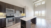 This single-floor space showcases a thoughtfully designed open-concept layout that emphasizes volume and light. The living area is particularly striking, featuring dramatically tall ceilings that rise well above standard height, lined with a generous arrangement of windows positioned at multiple levels. This design choice floods the space with natural light while offering outdoor views, with light hardwood flooring reflecting the brightness throughout.

The kitchen is a standout feature, outfitted with dark espresso cabinetry extending to ceiling height for maximum storage capacity. A full suite of stainless steel appliances includes a refrigerator, range, microwave, and dishwasher. Light quartz countertops provide strong contrast against the cabinetry, complemented by a subway tile backsplash with under-cabinet lighting. A central island with open shelving below serves as both a functional workspace and a visual anchor connecting the kitchen to the living area.

The bathroom is well-appointed with large-format tile flooring, a double-sink vanity featuring dark cabinetry with brushed nickel hardware, a wide mirror with overhead lighting, and a separate soaking tub and shower area. Natural light enters through a window, enhancing the clean, neutral palette that carries throughout the entire space.
