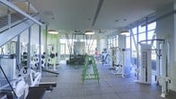 This two-floor fitness facility is designed to accommodate a comprehensive range of workout needs in a spacious, well-lit environment. The main workout floor is expansive, featuring rubber flooring throughout for durability and safety. Large floor-to-ceiling windows line the perimeter, flooding the space with abundant natural light and offering open views of the surrounding exterior.

The layout is thoughtfully organized, with distinct zones for various types of training. A dedicated free weights area includes a full rack of dumbbells arranged on tiered stands, complemented by barbells and weight plates on nearby racks. Resistance cable machines and multi-station strength equipment are positioned along one side, providing options for targeted muscle training. Green-accented equipment and wall features add a vibrant, energetic aesthetic throughout the space.

Overhead, ceiling fans and pendant lighting fixtures are suspended from the high ceilings, contributing to both airflow and illumination across the workout floor. Mirrors are strategically placed to enhance the sense of depth. The overall ambiance is modern and motivating, combining functional equipment arrangement with an open, airy atmosphere suited for a variety of fitness routines.