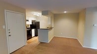 This single-floor space showcases an open-concept design that seamlessly connects the kitchen and living area. The kitchen is equipped with black appliances, including a refrigerator and range, complemented by white cabinetry and dark countertops. A peninsula with a dark countertop extends into the living space, creating a natural divide while maintaining an open, connected feel. Track lighting illuminates the kitchen, while recessed lighting adds brightness throughout. The kitchen features hardwood-style flooring, which transitions into neutral carpeting in the living area. The walls are finished in a warm beige tone, contributing to a clean and cohesive aesthetic. White baseboards provide a crisp contrast along the perimeter of the space. The layout is practical and efficient, offering a comfortable flow between the cooking and living zones.