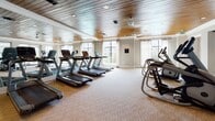 This fitness center is thoughtfully designed to offer a premium workout experience within an elegantly styled single-floor facility. The main exercise area is generously proportioned, accommodating a full suite of cardio equipment including multiple treadmills and elliptical machines arranged across neutral-toned, textured carpeting. Warm wood-plank ceilings with recessed lighting lend a refined, upscale character to the space, while floor-to-ceiling mirrors along one wall visually expand the room and enhance natural light. Large windows frame views of the surrounding greenery, creating an airy, open atmosphere that encourages an energizing workout environment. A ceiling fan provides additional comfort during exercise sessions. Adjacent to the main workout floor is a secondary zone, partitioned by expansive glass panels, which offers additional equipment and maintains visual connectivity throughout the space. A dedicated amenity nook provides practical conveniences, including a compact refrigerator stocked with beverages and paper towel dispensers for post-workout cleanup. A separate lounge area furnished with seating and a wall-mounted television offers a comfortable space to cool down or unwind. The overall design balances functionality with sophisticated interior finishes, making this fitness center both practical and visually inviting.