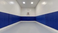 This single-level indoor basketball court is purpose-built for athletic training and recreational play. The space is defined by its clean, functional design, with white upper walls and bold blue padded wall mats lining the lower perimeter on all sides. These padded panels serve as both a safety feature and a visual design element, providing player protection during active gameplay while contributing to the court's sharp, two-tone aesthetic.

The court features a wall-mounted clear acrylic backboard with a standard orange rim and net, positioned at regulation height on the end wall. The smooth, light grey sport flooring spans the full area of the court, offering a consistent and durable playing surface well-suited for basketball drills, shooting practice, and general athletic use.

Overhead, recessed flush-panel ceiling lights provide even, bright illumination throughout the space, ensuring excellent visibility across the entire court. A standard access door is integrated into the padded rear wall, maintaining the seamless look of the court's interior. The enclosed design and high ceiling clearance create an immersive, distraction-free environment suitable for focused athletic training or casual recreational use.