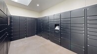 The Glenbrook Apartments Package Center in Cupertino, California is a purpose-built, single-room facility designed to streamline package delivery and retrieval for residents. The space features a clean, functional layout with neutral-toned walls and light polished concrete flooring that contributes to a bright, utilitarian aesthetic.

Dominating the room are banks of Luxer One smart package lockers finished in a sleek dark graphite, arranged along multiple walls in an L-shaped and parallel configuration. The lockers vary in size, accommodating parcels of different dimensions, and are equipped with a central digital kiosk for automated access and notifications. Posted signage directs carriers to use the lockers rather than leaving packages unattended, underscoring the facility's emphasis on secure delivery practices.

Natural light filters into the space through a skylight overhead and a glass-paneled exterior door, complementing the recessed ceiling lighting and lending the room an open, airy feel despite its compact footprint. A security camera is mounted near the ceiling, providing surveillance coverage of the entire room. The exterior entrance offers a view of surrounding greenery, integrating a subtle connection to the outdoor landscaping of the Cupertino community. Overall, the Package Center is a well-organized, modern amenity that prioritizes security, accessibility, and ease of use.