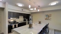 This single-floor residence showcases a thoughtfully designed interior with a strong emphasis on modern aesthetics and functional living. The kitchen serves as the heart of the space, featuring rich dark cabinetry that contrasts elegantly against light-toned walls, creating a bold and sophisticated look. Stainless steel appliances, including a refrigerator, dishwasher, and gas range, provide a sleek, professional-grade cooking environment. A deep basin sink and ample counter space further enhance the kitchen's practicality. The expansive breakfast bar, finished in a light granite-style surface, extends generously to accommodate multiple bar stools, making it an ideal spot for casual dining and entertaining. Overhead, a combination of flush-mount ceiling fixtures and a striking track lighting system with elongated pendant lights illuminates the space with layered, warm lighting. The neutral wall tones throughout create a clean, versatile backdrop that complements the dark cabinetry and bright countertops. The open layout between the kitchen and adjacent areas promotes a seamless flow, contributing to an airy and welcoming ambiance throughout this well-appointed residence.