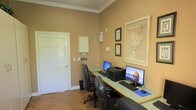 This single-story space includes a well-appointed workstation area designed for productivity and functionality. The room features warm, tan-toned walls accented by white crown molding, lending a clean and polished aesthetic. The flooring is laid with earthy stone tiles that complement the overall warm color palette.

The workstation is set up along an L-shaped or linear desk surface, accommodating two computer monitors and a printer, making it suitable for multi-user or multi-task use. Two black leather ergonomic office chairs provide comfortable seating at the stations. A white panel board is mounted on the wall near the entrance door, likely serving as a notice board or communication area. Electrical and utility access points are neatly integrated into the wall nearby.

Decorative wall art, including a prominently framed classical urn print and two smaller framed pieces, adds an elegant touch to the functional workspace. The white paneled door provides access to adjoining areas. The overall ambiance balances practicality with refined décor, making it a comfortable and organized environment for work or business-related activities.
