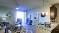 This single-floor space showcases an open-concept design that integrates the kitchen, dining, and living areas into one cohesive, flowing layout. The kitchen features granite-style countertops and is equipped with a pendant light fixture, adding a stylish focal point above the dining area. The living space is generously sized, highlighted by wood-style flooring and a ceiling fan that enhances both comfort and aesthetic appeal. A colorful area rug anchors the seating area, which is furnished with a classic accent chair and a glass-topped console table. Large sliding glass doors flood the space with natural light and provide direct access to an outdoor area, creating a bright and airy atmosphere. A doorway off the main living area leads into a bedroom, visible in the background, which appears well-lit and comfortably appointed with coordinated bedding and furnishings. The overall ambiance is modern yet inviting, with neutral wall tones complementing the warm wood flooring throughout. The open sightlines between rooms enhance the sense of space, making this a functional and visually connected living environment.