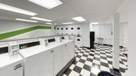 This communal laundry facility is designed for practicality and ease of use, offering a clean, well-organized environment for residents. The space is immediately recognizable by its bold black-and-white checkered tile flooring, which lends a classic, timeless aesthetic throughout the room. Bright, recessed panel lighting in the drop ceiling ensures the entire area is evenly and thoroughly illuminated.

The main laundry area is equipped with a row of top-load washing machines along one side, complemented by a generous bank of stacked dryers lining the opposite wall, providing ample capacity for multiple users at once. A folding table is conveniently positioned near the entrance, offering a dedicated surface for sorting and folding laundry. A payment kiosk is also present, facilitating easy access to machine operation.

A designated waiting area features white molded seating along a bench, set against a wall accented with a vibrant green horizontal stripe, adding a pop of color to the otherwise neutral palette. A wall clock and small decorative touches complete the seating nook.

Natural light filters in through a glazed exterior door, which also provides direct outdoor access. The space is further supported by a utility sink and a laundry cart for added convenience, making this facility a fully functional and user-friendly amenity.