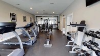 The Spring Creek fitness center is a thoughtfully designed, single-floor exercise facility offering a comprehensive workout environment for residents and guests. The gym features durable rubberized flooring throughout, providing both safety and functionality for a wide range of exercises. A variety of cardio equipment lines the space, including treadmills and elliptical machines, while a multi-station cable and weight machine anchors the strength training area. A dedicated free weights section with a flat bench allows for versatile strength workouts. A large wall-mounted mirror at the far end of the room visually expands the space and allows users to monitor their form. Recessed lighting and a ceiling fan ensure the room remains well-lit and comfortably ventilated. The neutral, light-toned walls paired with framed motivational artwork contribute to an energizing yet clean aesthetic. A water station is conveniently available within the space. The layout is open and well-organized, making efficient use of the available floor space while maintaining clear pathways between equipment. Overall, the facility provides a fully functional and welcoming environment for achieving a wide range of fitness goals.