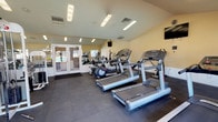 This fitness center is a thoughtfully designed, single-floor facility that offers a comprehensive workout environment for a wide range of exercise needs. The space features a vaulted ceiling that lends an open, airy feel, complemented by abundant natural light streaming through large windows that line the walls and offer views of the surrounding outdoor area, including an adjacent pool. Recessed fluorescent lighting ensures the interior remains bright throughout.

The flooring throughout is covered in durable, dark rubber interlocking tiles, providing safety and comfort during exercise. Full-length wall mirrors are installed along multiple walls, visually expanding the space and allowing users to monitor their form during workouts.

The cardio section is well-stocked with multiple treadmills, elliptical machines, and stationary bikes arranged in an orderly fashion. The strength-training area features a variety of cable and weight machines designed to target different muscle groups, including cable pulleys, lat pull-down stations, seated row machines, and a functional trainer. A flat bench is also available for free-weight exercises.

A set of glass double doors provides access directly to the outdoor pool area, enhancing the facility's connectivity to the broader recreational amenities. The overall atmosphere is clean, organized, and functional, making it a practical and inviting space for daily fitness use.