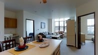 This single-floor residence offers an open-concept design that seamlessly integrates the kitchen, dining, and living areas into one cohesive space. The kitchen features a generous counter that doubles as a casual dining surface, accompanied by seating and ample workspace. Neutral-toned cabinetry and a full-size refrigerator contribute to the functional layout.

The dining area sits adjacent to the kitchen, accommodating a small table and chairs, making it well-suited for everyday meals. The living room extends naturally from this space, furnished with a plush sofa, an armchair, and a central coffee table. A ceiling fan overhead provides both ventilation and ambient lighting.

Large grid-pane windows along the far wall flood the living area with natural light and offer views of the surrounding neighborhood. The light-colored walls and carpeted flooring create a clean, neutral atmosphere throughout. A doorway visible from the main living area leads to an additional room, suggesting further private spaces beyond the open layout. Overall, the unit presents a practical and comfortable living environment with a straightforward, functional design.