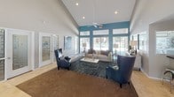 This single-story community clubhouse or leasing office is designed to be both functional and welcoming. The centerpiece of the space is a dramatic vaulted ceiling that soars overhead in the main lounge area, creating an open and airy atmosphere. A ceiling fan circulates air throughout the room, which is bathed in natural light streaming through large glass doors and transom windows framed in a bold teal accent wall. This vibrant color detail adds a modern, contemporary touch that carries consistently throughout the interior.

The main lounge area is comfortably appointed with a neutral sofa, leather accent chairs, a patterned area rug, and a glass coffee table, making it an inviting space for guests. Adjacent to the lounge, a dedicated workstation with a dark desk and seating serves as the primary reception or leasing area. Decorative artwork adorns the walls, further enhancing the professional yet comfortable environment.

Large glass doors open directly to an outdoor pool area, seamlessly connecting the interior with outdoor amenities. Tile flooring runs throughout the space, adding to the clean, polished aesthetic.

The kitchenette area features dark granite countertops, light wood cabinetry, a microwave, a sink, and a wall-mounted flat-screen TV, providing a well-equipped refreshment station for staff and visitors alike. Frosted glass windows branded with the property logo lend a refined, professional finish to this area.