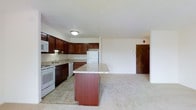 This single-floor residence offers an open and functional layout that seamlessly connects the kitchen and living areas. The kitchen is a standout feature, appointed with rich dark wood cabinetry that extends to the upper and lower sections of the walls, providing generous storage throughout. Light granite countertops run along the perimeter and are echoed on the central kitchen island, which serves as a natural gathering point and additional workspace. White appliances, including a range, refrigerator, and built-in microwave, complement the clean, neutral palette of the space. A stainless steel sink is positioned beneath the cabinetry, and tile flooring grounds the kitchen zone with a practical, easy-to-maintain surface. The kitchen transitions smoothly into the open living area, where plush carpeting creates a softer, more comfortable atmosphere. The space is bathed in a bright, airy ambiance, enhanced by light-toned walls and flush ceiling lighting. A solid entry door is visible at the far end of the living area, lending a sense of boundary and privacy to the overall layout. The combination of warm wood tones, light stone surfaces, and open sightlines gives this residence a welcoming and well-organized character.