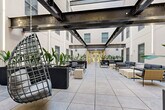 This courtyard is a thoughtfully designed semi-enclosed outdoor gathering space set within the footprint of a multi-story building, open to the sky above while surrounded by the structure's white-painted facades and grid-patterned windows on multiple levels. One of its most defining architectural features is the network of large, exposed industrial steel beams that span the width of the space overhead, adding a bold, raw contrast to the otherwise clean and contemporary aesthetic.

The flooring throughout consists of large-format light gray tiles laid in a precise grid pattern, contributing to the spacious and ordered feel of the courtyard. Generous open floor space allows for flexible arrangement of seating zones. Multiple distinct areas are furnished for both casual lounging and social dining: modular sofas and low-profile chairs with cushioned seating are arranged in conversation groupings, while a separate dining area features dark metal tables paired with olive green chairs. Hanging woven egg chairs suspended by chains add a playful, decorative element.

Tall planters filled with tropical foliage, including bird-of-paradise plants, are positioned throughout the space, introducing lush greenery that softens the industrial surroundings. Glass double doors at one end provide interior access, completing this versatile and visually engaging communal courtyard.