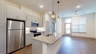 This contemporary single-floor unit is designed with a clean, modern aesthetic and a practical open-concept layout. The kitchen serves as the centerpiece of the living space, showcasing a large granite-topped island with an undermount sink and a professional-style pull-down faucet. Soft gray shaker cabinets line the walls, complemented by a white tile backsplash and a full suite of stainless steel appliances, including a refrigerator, electric range, and built-in microwave. Two geometric gold pendant lights hang above the island, adding a stylish focal point. The kitchen flows seamlessly into the living area, which features warm hardwood-style flooring, neutral white walls, and a ceiling fan. A glass door provides access to an outdoor area, allowing natural light to fill the space. A built-in desk and shelving nook off the kitchen offers a dedicated workspace. The bedrooms are bright and well-proportioned, each with large windows that frame outdoor views and continue the hardwood-style flooring. The primary suite includes a generously sized walk-in closet outfitted with multiple shelving tiers and hanging rods. The bathroom features granite countertops, gray cabinetry with brushed nickel hardware, an undermount sink, and a large framed mirror, maintaining the cohesive, modern finish found throughout the unit.