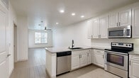 This thoughtfully designed single-floor residence offers a cohesive, modern living experience across a comfortable layout encompassing one bedroom, one bathroom, a kitchen, living room, and walk-in closet. Light-toned wood-look tile flooring runs throughout the main living areas, creating a clean and cohesive aesthetic, while plush carpet adds warmth to the bedroom and closet spaces.

The kitchen is a standout feature, outfitted with white shaker-style cabinetry, granite-look countertops, a peninsula with seating potential, and a full suite of stainless steel appliances including a refrigerator, range, over-the-range microwave, and dishwasher. A deep single-basin sink with a matte black faucet anchors the space. The kitchen flows seamlessly into the living area, which is brightened by a large window with outdoor views and finished with a ceiling fan overhead.

The bedroom offers a serene retreat with plush carpet, a ceiling fan, and a window that invites natural light. It connects directly to a generously sized walk-in closet with wire shelving and to a well-appointed bathroom featuring white cabinetry, a wide vanity with a framed mirror, matte black fixtures, tile flooring, and a soaking tub with a tiled surround. A stacked in-unit washer and dryer add exceptional convenience to this well-rounded residence.