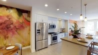This single-floor space combines modern design with artistic flair, creating an inviting and visually dynamic environment. The kitchen is a standout feature, outfitted with a full suite of stainless steel appliances, including a French door refrigerator with an in-door ice and water dispenser, a gas range with oven, an over-the-range microwave, and a dishwasher. Crisp white upper and lower cabinetry provides generous storage, while a classic white tile backsplash adds texture and clean contrast. Light wood-toned flooring runs throughout, contributing to a bright and airy atmosphere enhanced by recessed ceiling lighting. A countertop peninsula extends the workspace and defines the kitchen area, offering additional surface space for dining or food preparation. What truly distinguishes this space is the bold, large-scale watercolor-style wall murals that flank the kitchen — rich with warm tones of coral, peach, and pink — infusing the interior with a striking artistic character. Complementary pops of orange accent walls and lush greenery from potted plants further enrich the space's vibrant, curated aesthetic, making it as visually expressive as it is functional.