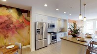 This single-floor space combines modern design with artistic flair, creating an inviting and visually dynamic environment. The kitchen is a standout feature, outfitted with a full suite of stainless steel appliances, including a French door refrigerator with an in-door ice and water dispenser, a gas range with oven, an over-the-range microwave, and a dishwasher. Crisp white upper and lower cabinetry provides generous storage, while a classic white tile backsplash adds texture and clean contrast. Light wood-toned flooring runs throughout, contributing to a bright and airy atmosphere enhanced by recessed ceiling lighting. A countertop peninsula extends the workspace and defines the kitchen area, offering additional surface space for dining or food preparation. What truly distinguishes this space is the bold, large-scale watercolor-style wall murals that flank the kitchen — rich with warm tones of coral, peach, and pink — infusing the interior with a striking artistic character. Complementary pops of orange accent walls and lush greenery from potted plants further enrich the space's vibrant, curated aesthetic, making it as visually expressive as it is functional.