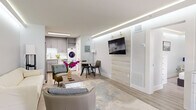 This thoughtfully designed single-floor unit maximizes every square foot through an open-concept layout and cohesive modern aesthetic. A signature design element found throughout the space is the recessed LED cove lighting along the ceiling perimeter, casting a clean, ambient glow across each room.

The living area is open and airy, featuring light hardwood-style flooring, neutral walls, and a wall-mounted television anchored by a wood-panel accent wall. A seating arrangement with a cream sofa and accent chair surrounds a white coffee table, creating a comfortable gathering space. Adjacent to the living area, a compact dining nook with a round table connects seamlessly to the fully equipped kitchen.

The kitchen features gray shaker-style cabinetry, stainless steel appliances including a refrigerator, range, microwave, and dishwasher, complemented by a marble-look tile backsplash and white quartz countertops. A stacked washer and dryer unit is conveniently integrated nearby.

The bedroom offers a tranquil retreat with the same cove lighting detail, a low-profile window with roller shades, and mirrored closet doors. The bathroom is finished with marble-look tile surrounds, a frameless glass shower enclosure with matte black hardware, and gray vanity cabinetry matching the kitchen.