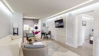 This thoughtfully designed single-floor unit maximizes every square foot through an open-concept layout and cohesive modern aesthetic. A signature design element found throughout the space is the recessed LED cove lighting along the ceiling perimeter, casting a clean, ambient glow across each room.

The living area is open and airy, featuring light hardwood-style flooring, neutral walls, and a wall-mounted television anchored by a wood-panel accent wall. A seating arrangement with a cream sofa and accent chair surrounds a white coffee table, creating a comfortable gathering space. Adjacent to the living area, a compact dining nook with a round table connects seamlessly to the fully equipped kitchen.

The kitchen features gray shaker-style cabinetry, stainless steel appliances including a refrigerator, range, microwave, and dishwasher, complemented by a marble-look tile backsplash and white quartz countertops. A stacked washer and dryer unit is conveniently integrated nearby.

The bedroom offers a tranquil retreat with the same cove lighting detail, a low-profile window with roller shades, and mirrored closet doors. The bathroom is finished with marble-look tile surrounds, a frameless glass shower enclosure with matte black hardware, and gray vanity cabinetry matching the kitchen.