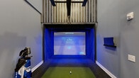 Located in Columbus, Ohio, this dedicated indoor golf simulator suite delivers a premium experience in a thoughtfully designed space. The room features soaring ceilings that accommodate a Full Swing golf simulator, complete with a ceiling-mounted projector, a large impact screen framed by vivid blue LED lighting, and a synthetic turf hitting mat. A wood-slat accent panel above the simulator bay adds a refined architectural touch, while the ceiling-mounted swing tracking arm ensures accurate shot data.

The lounge area is furnished with rich cognac leather armchairs arranged on a woven area rug over warm wood-look plank flooring, creating a comfortable space for guests to relax between rounds. A dramatic floor-to-ceiling black-and-white golf course mural anchors the lounge wall, complemented by framed golf-themed artwork throughout. A wall-mounted flat-screen television and a ceiling-mounted secondary projector provide additional viewing options.

A built-in wet bar with dark cabinetry, under-counter beverage refrigeration, and bar seating lines one wall, making the space well-suited for entertaining. A mini-split HVAC unit ensures climate comfort year-round. The overall design strikes a balance between sporty functionality and upscale lounge aesthetics, making this suite an ideal venue for practice sessions, competitive play, or social gatherings centered around golf.