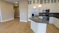 This single-floor space showcases a thoughtfully designed interior that seamlessly blends the sleeping and living areas through the use of sleek, industrial-style sliding barn doors. The doors, fitted with frosted glass panels and mounted on exposed metal hardware tracks, provide both privacy and an architectural focal point, allowing the bedroom to be opened to or separated from the main living area as needed.

The bedroom alcove features a well-appointed bed dressed in neutral, layered bedding with accent pillows, complemented by wall-mounted sconce lighting and decorative diamond-shaped wall art. A bold, dark navy accent wall in the living area creates a dramatic contrast against the lighter gray walls, adding depth and visual interest to the overall design.

The living space is furnished with a curved, cream-upholstered accent chair with nailhead trim detailing, a plush ottoman, and a classic floor lamp with a white shade. A patterned area rug anchors the seating arrangement, adding warmth and texture to the dark hardwood flooring. The combination of materials, tones, and layered lighting gives the space a sophisticated yet comfortable ambiance.