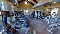 This fitness center offers a thoughtfully designed workout environment housed within an architecturally distinctive single-floor space. The room is anchored by soaring vaulted ceilings with exposed wooden beams, lending a warm and open atmosphere that sets it apart from a typical gym setting. A stylish chandelier and recessed lighting illuminate the space, complementing the natural light that streams in through multiple windows and French doors along the walls. Full-length mirrors line one side of the room, visually expanding the space and allowing users to monitor their form during exercise. The flooring features durable, cushioned carpet suited for a high-traffic fitness environment. The cardio section is well-stocked with elliptical trainers, treadmills, and a rowing machine, while the strength training area offers a variety of cable and resistance machines to support a comprehensive full-body workout. Wall-mounted flat-screen televisions are positioned for easy viewing from multiple points in the room, enhancing the workout experience. The combination of functional equipment, airy architecture, and natural light creates an inviting and motivating space for fitness enthusiasts of all levels.