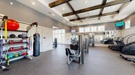 Located at 1300 S Davis Rd in LaGrange, Georgia, this generously sized fitness center offers a thoughtfully designed, single-floor layout that accommodates a full range of workout activities. The space immediately impresses with its soaring ceilings adorned with distinctive rustic dark wood box beams, creating an inviting contrast against the crisp white ceiling and soft grey walls. Recessed lighting and ceiling fans are distributed throughout, ensuring excellent visibility and comfort during workouts.

Large multi-pane windows line the walls at both standard and clerestory heights, flooding the interior with natural light and offering views of the surrounding greenery. Durable speckled rubber flooring covers the entire workout area, providing both safety and functionality.

The fitness center is comprehensively equipped, featuring a dedicated strength training zone with cable machines, weight benches, and a full dumbbell rack, complemented by a floor-to-ceiling mirror wall that enhances the sense of space. A cardio section houses treadmills, stationary spin bikes with interactive screens, and stair-climbing machines. A boxing area with a hanging heavy bag adds variety for high-intensity training. Wall-mounted televisions are positioned for convenient viewing throughout. The overall ambiance is bright, modern, and energizing.