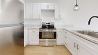 This 670 sq ft, single-floor apartment at 1 Noelle Court in Amityville, NY offers 2 bedrooms and 1 bathroom. The open kitchen-living area features white shaker cabinets, marble-style countertops, stainless steel appliances including a range, refrigerator, and dishwasher, plus pendant lighting. Light hardwood floors and white walls run throughout, creating a bright, clean aesthetic. The living room includes recessed lighting, baseboard heating, and a wall A/C unit. Bedrooms offer natural light via windows with green outdoor views, closets, and baseboard heating. The bathroom includes a vanity and tub/shower combo.