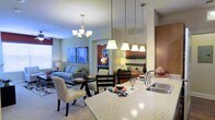 Located at 320 The Green Cir in Raleigh, NC, this single-story residence showcases an open-concept design that integrates the kitchen, dining, and living areas into one cohesive and functional space. The kitchen is appointed with granite countertops, a stainless steel sink, and a series of pendant lights suspended above the island, providing both task lighting and visual appeal. The dining area sits centrally within the open layout and is illuminated by an elegant chandelier, creating a defined gathering space without physical separation from the surrounding rooms. The living room offers a relaxed atmosphere, furnished with a comfortable sofa and seating arrangement set atop a patterned area rug. Large windows allow natural light to fill the space, complemented by a ceiling fan for added comfort. The lofty ceilings contribute to an airy, spacious feel throughout. A glimpse into an adjacent bedroom reveals warm accent walls and coordinated décor, maintaining a consistent design aesthetic. Overall, the space presents a well-lit, modern interior with thoughtful architectural details and a welcoming ambiance throughout.