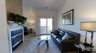 This single-floor space centers around a well-appointed living room that balances comfort and contemporary style. The room features elevated ceilings that lend an open, airy feel, enhanced by neutral white walls that reflect natural light streaming in through a large sliding glass door. The door leads to an outdoor balcony, offering views of surrounding greenery and bringing a connection to the outdoors into the living space.

The living area is anchored by a tufted dark leather sofa accented with patterned throw pillows, paired with a sleek glass-top coffee table on a plush area rug over carpeted flooring. A matching leather armchair is positioned near the fireplace, which features a tile surround and serves as a focal point on one side of the room. A framed piece of wall art adds a decorative touch above the sofa, contributing to the polished aesthetic. A small table lamp provides warm ambient lighting to complement the natural light. The overall ambiance is modern and inviting, with a cohesive design that makes effective use of the available space.