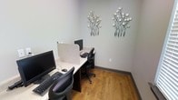 This single-floor space is a compact yet functional computer room designed to facilitate focused, individual work. The room is outfitted with a shared workstation desk that runs along the wall, divided into separate stations by privacy panels, each equipped with a desktop computer, monitor, keyboard, and mouse. The ergonomic mesh-back chairs at each station offer comfort during extended use. The warm hardwood-style flooring adds a welcoming touch to the otherwise utilitarian setup, grounding the space with a sense of warmth and approachability. The walls are painted in a soft, neutral gray-green tone that promotes a calm and productive atmosphere. Two decorative metal wall sculptures featuring branching tree motifs with circular accents add an artistic flair, softening the functional nature of the room. The layout is efficient and intimate, making it well-suited for tasks requiring concentration. Overall, the space balances practicality with thoughtful design touches, creating an environment that is both purposeful and visually pleasant — ideal for use as a shared business center or resource room within a larger facility.