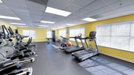 This fitness center offers a comprehensive workout environment designed to support a wide range of exercise routines. The space is finished with durable rubber tile flooring, providing both safety and comfort during training sessions. Bright yellow accent walls energize the atmosphere, while multiple windows along the perimeter flood the room with natural light, creating an open and inviting ambiance. The drop ceiling is fitted with recessed fluorescent panel lighting, ensuring consistent illumination throughout. The cardio area is well-appointed with multiple treadmills and a stationary bike, catering to various cardiovascular training preferences. A dedicated strength training zone features a multi-station cable machine, a dumbbell rack stocked with free weights, and stability equipment including an exercise ball. The layout is thoughtfully organized, allowing ample space between equipment for safe and comfortable use. The overall design balances functionality with an uplifting aesthetic, making it a practical and motivating facility for daily fitness routines.