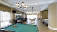 This single-floor amenity lounge is thoughtfully designed to serve as both a social hub and a relaxed gathering space. The open-concept layout seamlessly connects several distinct activity zones, creating an atmosphere that is equally suited for casual interaction and structured socializing.

At the forefront of the space sits a full-size billiards table with a classic green felt surface, serving as a natural focal point and centerpiece for recreation. Overhead, a contemporary multi-arm chandelier with gold-toned cylindrical shades adds a touch of elegance and provides warm, ambient lighting.

Adjacent to the billiards area, a bar-height counter with sleek dark surfaces and gold-accented stools offers a stylish spot for casual seating. A wall-mounted television nearby enhances the entertainment options available in this zone.

Toward the back of the space, a communal high-top table surrounded by upholstered barrel chairs creates an ideal setting for group gatherings or informal dining. Pendant lighting above this area adds an intimate, layered lighting effect.

Large windows along the rear wall flood the space with natural light and contribute to the bright, airy feel of the room. The warm taupe wall tones, light wood-look flooring, and lush potted greenery tie the entire space together, creating a polished yet welcoming environment.