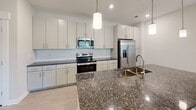 Located at 4212 Napa Oak Dr in Charlotte, NC, this single-story home offers a thoughtfully designed layout with modern finishes throughout. The open-concept kitchen serves as the centerpiece of the living area, showcasing dark granite countertops, white shaker-style cabinetry, a subway tile backsplash, and a full suite of stainless steel appliances including a range, microwave, and French door refrigerator. A large central island with a double-basin sink and pendant lighting above provides both additional prep space and a natural gathering point. Light wood-tone flooring runs through the main living and kitchen areas, while plush carpeting adds warmth to the bedrooms. The bedrooms are well-proportioned, each featuring neutral tones and windows that allow natural light and outdoor views of surrounding greenery. One bedroom offers direct exterior access through a glass-paneled door flanked by wide windows. The bathrooms are finished with granite vanity tops, undermount sinks, brushed nickel fixtures, and large framed mirrors. A full bathroom includes a soaking tub and shower combination with clean white tile surround. Throughout the home, clean lines, recessed lighting, and a neutral color palette create a cohesive, move-in-ready aesthetic.