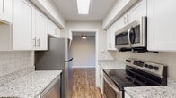 This single-floor residence offers a thoughtfully designed layout that balances comfort and functionality throughout its living spaces. The galley-style kitchen is a standout feature, showcasing granite countertops, crisp white cabinetry with brushed nickel hardware, and a full suite of stainless steel appliances including a refrigerator, electric range, and over-the-range microwave. A skylight overhead fills the kitchen with natural light, while wood-look plank flooring adds warmth underfoot and carries through into the main living areas. The open living and dining area is generously proportioned, featuring a ceiling fan and sliding glass doors that lead to a private balcony overlooking a lush, tree-lined outdoor setting. The bedrooms are finished with plush wall-to-wall carpeting, neutral-toned walls, and ample natural light provided by well-placed windows with blinds. The bathroom continues the refined aesthetic with granite countertops on white vanity cabinetry, wood-look flooring, and a tiled shower surround with a marble-style finish. Throughout the residence, consistent use of neutral tones, quality finishes, and practical design elements create a clean, modern ambiance that is both inviting and move-in ready.