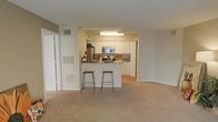 This single-floor residence offers a practical and comfortable open-concept layout that seamlessly connects the kitchen and living areas. The kitchen is well-appointed with white cabinetry, granite-style countertops, and stainless steel appliances including a refrigerator, microwave, and range. A peninsula counter extends into the living space, accompanied by two bar stools that create a convenient casual dining or breakfast bar area. The kitchen is illuminated by a flush overhead light fixture, ensuring a bright and functional workspace. The transition from the kitchen to the living area is smooth, with the flooring shifting from hard surface to plush wall-to-wall carpeting that adds warmth and comfort underfoot. The neutral color palette throughout — soft beige walls, white trim, and light carpet — creates a clean, cohesive aesthetic that is easy to personalize. Ventilation is well-considered, with visible HVAC vents integrated into the walls. The layout offers an efficient use of space, with clear sightlines from the living area into the kitchen, making it ideal for everyday living and casual entertaining. The overall ambiance is bright, understated, and move-in ready.