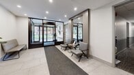 Located at 65 Paisley Blvd W in Mississauga, Ontario, this ground-floor lobby serves as a polished and functional gateway to the building. The space is designed with a contemporary aesthetic, featuring large-format light-toned floor tiles that run throughout, complemented by recessed pot lighting that ensures the area is consistently bright and inviting.

The entrance features full-height glass doors framed in dark metal, allowing natural light to filter into the lobby and providing clear sightlines to the exterior. A charcoal runner mat extends from the entrance inward, delineating the main circulation path. Adjacent to the entry, a cushioned bench provides a convenient waiting area, while a pair of modern grey lounge chairs arranged around a marble-topped accent table offer a more refined seating option, accented by warm wood-toned wall paneling.

Toward the rear of the lobby, two stainless steel elevator doors with polished finishes are prominently positioned, accompanied by clearly marked floor directory and wayfinding signage. A lush fiddle-leaf fig plant in a square planter adds a natural, organic element that softens the otherwise clean, modern interior. Security is supported by visible ceiling-mounted surveillance cameras. The overall ambiance is professional, tidy, and welcoming, reflecting a well-maintained residential building entrance.