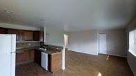 This single-story space is designed around an open-concept floor plan that seamlessly connects the kitchen and living area, creating a functional and cohesive environment. The kitchen is outfitted with dark wood cabinetry, stone-look countertops, and a tile backsplash that adds texture and visual interest. A peninsula with a built-in sink and dishwasher extends into the space, providing both additional workspace and a natural dividing point between the kitchen and the adjoining living area. A white support column anchors the peninsula, contributing to the architectural character of the layout. The living area is generously sized, offering ample room for various furniture arrangements. Hardwood-style flooring runs throughout, lending warmth and continuity across both areas. The walls are finished in a soft, neutral tone, complementing the overall palette and contributing to a clean, understated aesthetic. A doorway visible from the living area suggests access to additional rooms or utility spaces. The space benefits from recessed lighting and appears bright and well-maintained, making it a practical and inviting environment overall.