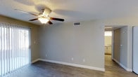 This single-story space presents a clean, functional layout designed for everyday living. The main living area features dark hardwood-style flooring that contrasts neatly with the light gray walls and crisp white baseboards, creating a polished, modern aesthetic. A five-blade ceiling fan with an integrated light fixture is centrally mounted, providing both air circulation and ambient lighting throughout the room.

The textured popcorn ceiling adds a classic architectural detail, while the HVAC vent positioned along the upper wall ensures efficient climate control. Electrical outlets are conveniently distributed along the walls, offering practical placement for furnishings and electronics.

A doorway visible along one wall provides a natural transition to an adjacent room, which appears to be a kitchen or utility area, suggesting an open and connected floor plan. The neutral color palette throughout creates a versatile backdrop, allowing for a wide range of interior design possibilities. Overall, the space offers a straightforward and functional environment with practical finishes well-suited for comfortable, day-to-day use.