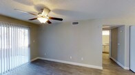This single-story space presents a clean, functional layout designed for everyday living. The main living area features dark hardwood-style flooring that contrasts neatly with the light gray walls and crisp white baseboards, creating a polished, modern aesthetic. A five-blade ceiling fan with an integrated light fixture is centrally mounted, providing both air circulation and ambient lighting throughout the room.

The textured popcorn ceiling adds a classic architectural detail, while the HVAC vent positioned along the upper wall ensures efficient climate control. Electrical outlets are conveniently distributed along the walls, offering practical placement for furnishings and electronics.

A doorway visible along one wall provides a natural transition to an adjacent room, which appears to be a kitchen or utility area, suggesting an open and connected floor plan. The neutral color palette throughout creates a versatile backdrop, allowing for a wide range of interior design possibilities. Overall, the space offers a straightforward and functional environment with practical finishes well-suited for comfortable, day-to-day use.