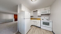 This single-floor unit at Glenora Gardens in Rochester, New York, offers a practical and well-organized layout across a compact footprint. The living room serves as the central gathering space, featuring neutral gray walls, wall-to-wall carpeting, and multiple windows that allow natural light to fill the room. Baseboard heating runs along the perimeter walls, and a wall-mounted air conditioning unit provides climate control year-round. The entry door connects directly to this open area, creating an accessible flow throughout the unit.

The kitchen is efficiently designed with white cabinetry extending to the ceiling for ample storage, warm wood-toned countertops, and light vinyl plank flooring. White appliances—including a full-size refrigerator, electric range, and over-the-range microwave—are neatly integrated into the layout. A hallway connects the main living areas to the private spaces beyond.

The bedroom offers a comfortable footprint with soft carpeting, neutral walls, a window with natural light, and a sliding double-door closet providing generous storage. A dedicated bathroom features classic tile-surround walls and a full soaking tub, maintaining a clean and functional aesthetic throughout the unit.