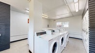 Located at 2770 Atlanta Ave in Riverside, California, this shared laundry facility offers a practical and well-organized space designed to serve its users efficiently. The room is bright and clean, with white-painted walls, fluorescent overhead lighting, and a durable concrete floor that ensures easy maintenance. Multiple front-loading washing machines are arranged in a row along the central floor space, while stacked dryer units line the back wall, maximizing the room's capacity. A granite-topped folding counter runs along one wall, providing a convenient surface for sorting and folding laundry. A distinctive dark slatted accent wall adds a modern design element, contrasting with the otherwise clean and neutral interior. The "met" branding is prominently displayed on one wall, reinforcing the facility's community identity. Windows on multiple walls allow natural light to filter in and provide views of the surrounding landscaping, including colorful flowers visible through the entry door. A fire extinguisher is mounted near the entrance for safety. The space is accessed through a windowed exterior door, offering a glimpse of the well-maintained grounds outside. Overall, the facility is thoughtfully equipped and maintained, providing residents with a clean, functional, and welcoming environment for their laundry needs.