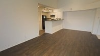 This single-floor space is defined by an open-concept layout that connects the living area and kitchen in a seamless, functional design. Dark hardwood flooring extends throughout, providing a rich, warm contrast against the crisp white walls and creating a cohesive visual flow across the space.

The living area is generously proportioned, offering an open and airy feel enhanced by high ceilings that add to the sense of volume. The clean, neutral walls provide a versatile backdrop suitable for a variety of interior styles.

The kitchen is partially separated from the living area by a white peninsula with a dark countertop, which doubles as a breakfast bar or prep surface. White cabinetry lines the kitchen walls, complemented by dark granite or stone countertops that add contrast and depth. Black appliances, including a refrigerator with freezer and a range with overhead ventilation, are integrated into the design. Track lighting mounted on the ceiling provides focused illumination throughout the kitchen area.

A doorway visible within the kitchen hints at additional rooms or storage space beyond. Overall, the space presents a clean, modern aesthetic with a practical, open layout.