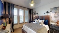 This single-floor residence offers a thoughtfully designed living environment with a focus on comfort and contemporary style. The bedroom serves as a standout feature, showcasing a rich brown leather upholstered headboard that anchors the space with a warm, sophisticated aesthetic. Soft neutral tones on the walls complement the plush carpeted flooring, creating an inviting and restful ambiance. The room is dressed with layered bedding in coordinating shades of grey, white, and navy, accented by an array of decorative pillows that add texture and visual depth. A row of double-hung windows fitted with horizontal blinds and floor-length striped drapes floods the room with natural light while offering views to the outdoors. Adjustable wall-mounted reading sconces flank the headboard, providing functional yet stylish task lighting, while a classic ceiling fan overhead ensures year-round comfort. A dark-toned nightstand with decorative accents completes the refined look. The overall atmosphere is one of curated elegance — balancing modern design sensibilities with cozy, residential warmth throughout the space.