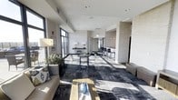 This single-floor communal space presents a sleek, modern aesthetic defined by clean lines, a neutral color palette, and thoughtfully curated furnishings. The open-plan layout is anchored by a bold, geometric patterned carpet in shades of charcoal and grey, which contrasts elegantly with the crisp white ceilings and walls. Recessed lighting is installed throughout, complemented by generous natural light streaming in through large, floor-to-ceiling windows that line one side of the space, creating a bright and welcoming atmosphere. The lounge area features a contemporary sofa adorned with decorative pillows, paired with a sleek coffee table, making it ideal for casual gatherings or relaxed conversation. Towards the rear of the space, bar-height seating is arranged along a counter area, offering a more upright setting for focused work or informal meetings. A textured stone accent column provides a striking architectural detail, adding depth and contrast to the otherwise smooth surfaces. Subtle decorative touches, including table lamps, small potted plants, and a glass vase, contribute warmth and personality to the environment. The overall ambiance is polished and professional, suited for use as a shared amenity or gathering area.