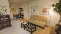 This single-floor space offers a warm and inviting atmosphere with a thoughtfully arranged open-concept layout that seamlessly connects the living and dining areas. The living area is anchored by a comfortable tan futon sofa, complemented by a pair of matching dark-framed side tables that add a modern, practical touch. Framed botanical and abstract artwork adorns the walls, lending the space a cultured and refined aesthetic.

A tall, dark ladder-style shelving unit stands near the entryway, providing both storage and display space while adding a touch of character to the room. The front door area is clean and accessible, creating a welcoming entry point into the home.

Transitioning toward the dining area, a dark wood table surrounded by matching chairs creates an intimate setting ideal for everyday meals or casual gatherings. The flooring transitions from plush carpet in the living zone to smooth hardwood near the dining and entry areas, adding visual interest and defining each functional space.

The overall ambiance is calm and neutral, with warm beige and earth-toned accents throughout. Recessed lighting provides a soft, even illumination across the space, enhancing its cozy and comfortable feel. This well-organized layout makes efficient use of the available space while maintaining a clean and livable environment.