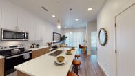 This single-floor residence showcases a thoughtfully designed open-concept layout that seamlessly connects the kitchen, dining, and living areas. The kitchen is a standout feature, equipped with stainless steel appliances, a combination of white upper cabinetry and warm wood-toned lower cabinets, white quartz countertops, and a generous island with an undermount sink and bar seating. Pendant lighting adds an elegant touch overhead, while recessed lighting and elevated ceilings enhance the sense of openness throughout.

The living area is anchored by a large sliding glass door dressed in vertical blinds, allowing abundant natural light to fill the space. Wood-look flooring runs throughout the main living areas, adding warmth and continuity. A dedicated dining nook sits adjacent to the living space, completing the open-plan arrangement.

The bedroom features plush carpeting, tall windows with horizontal blinds, and ample wall space. It connects directly to a well-designed bathroom appointed with a wood-toned vanity, white quartz countertop, a large mirror, and a walk-in shower. A walk-in closet with built-in rod storage provides generous organizational capacity. The overall aesthetic is clean, contemporary, and cohesive.